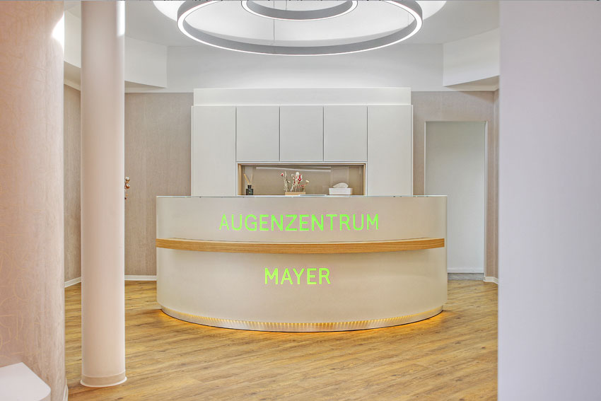 Empfangstresen steht im Raum, mit der Aufschrift "AUGENZENTRUM MAYER" in grün leuchtenden Buchstaben, umgeben von modernen, hellen Einrichtungsgegenständen und einem Holzfußboden.