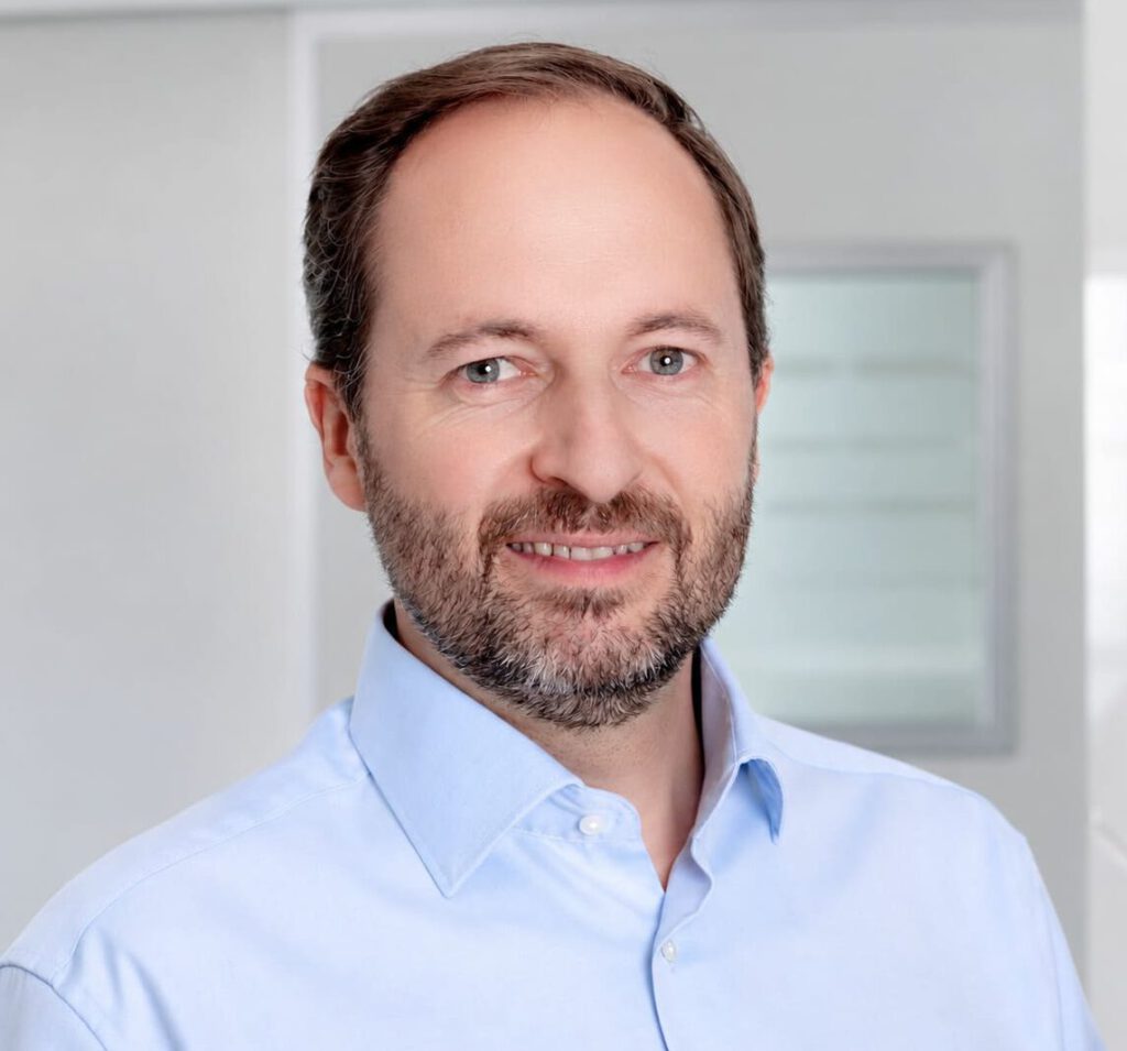 Dr. med. Max von Wedemeyer trägt ein blaues Hemd, lächelt freundlich und steht vor einem grauen Hintergrund.